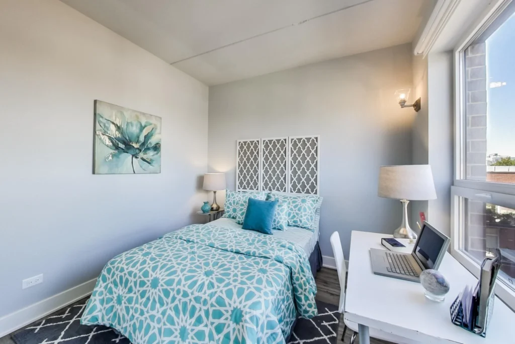 A neatly made bed with teal patterned bedding, a desk with a laptop and lamp, large window, and wall art in a bright, modern bedroom.