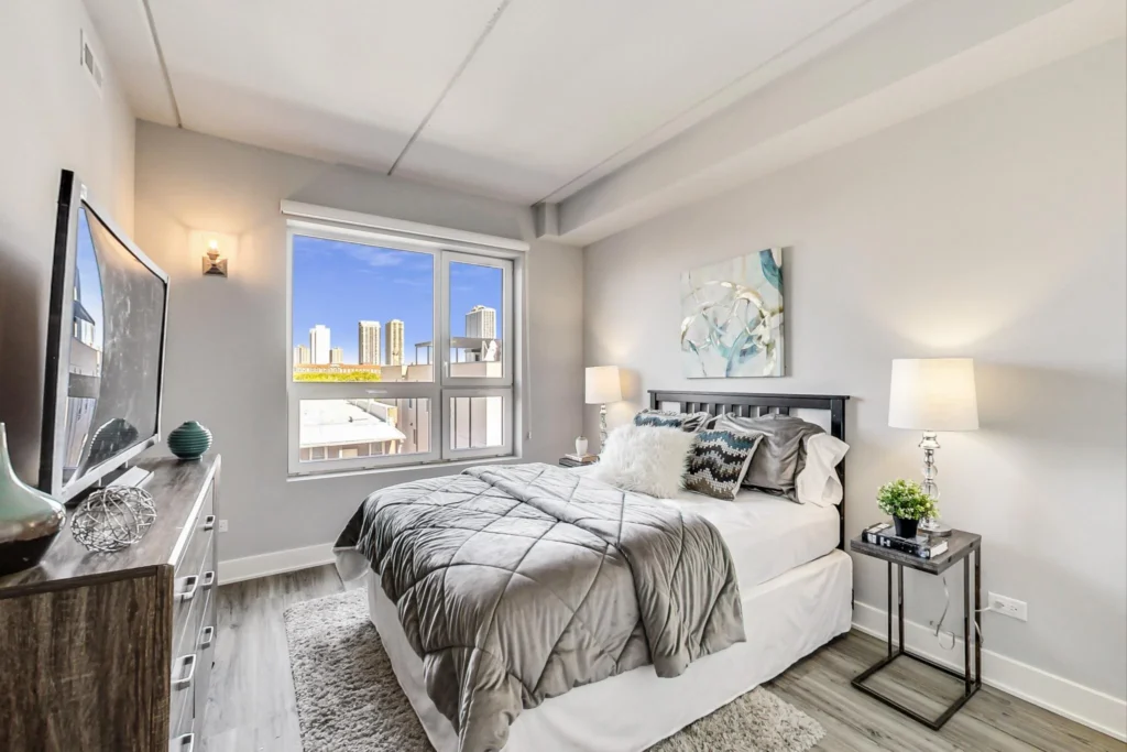 Modern bedroom with a neatly made bed, large window showing a city skyline, two bedside lamps, TV on a dresser, and light-colored decor.