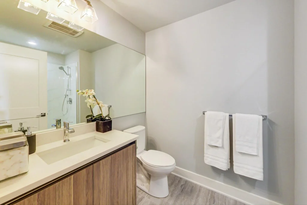 Modern bathroom with a large mirror, wooden vanity, white countertop, toilet, towel rack with two white towels, and a shower visible in the background.