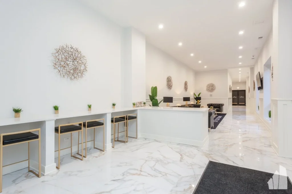 A modern lobby with white marble floors, a long counter with stools, potted plants, wall art, and recessed ceiling lights.