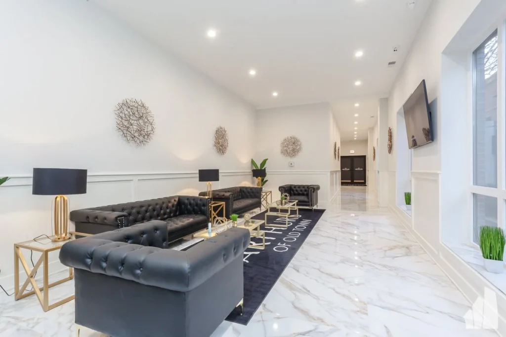 Modern lobby with black leather sofas, marble floors, wall art, two black lamps on gold tables, a large rug, and a mounted TV.