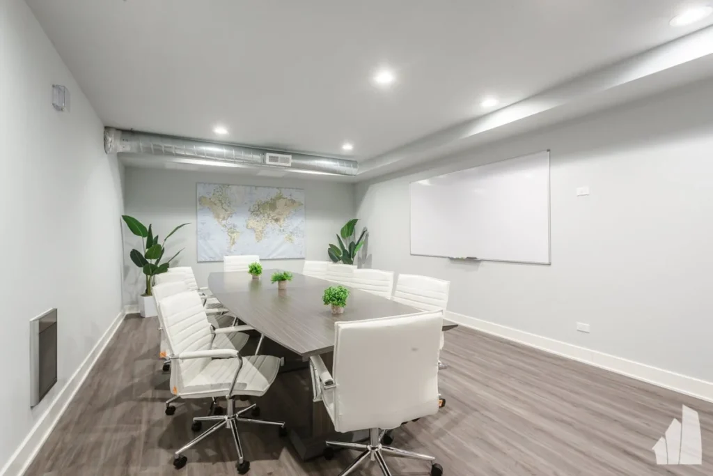 A modern conference room with a long table, white chairs, a large world map on the wall, plants in the corners, and a whiteboard.