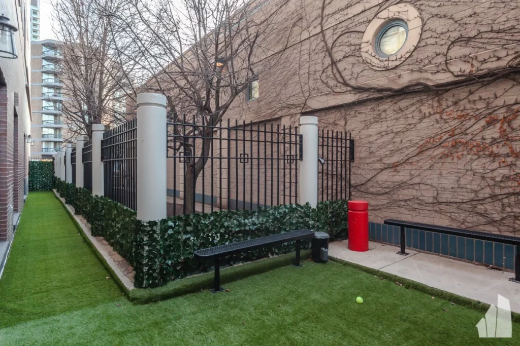 A narrow outdoor area with artificial grass, a black bench, a tennis ball, a red cylindrical bin, a vine-covered wall, and a black metal fence.