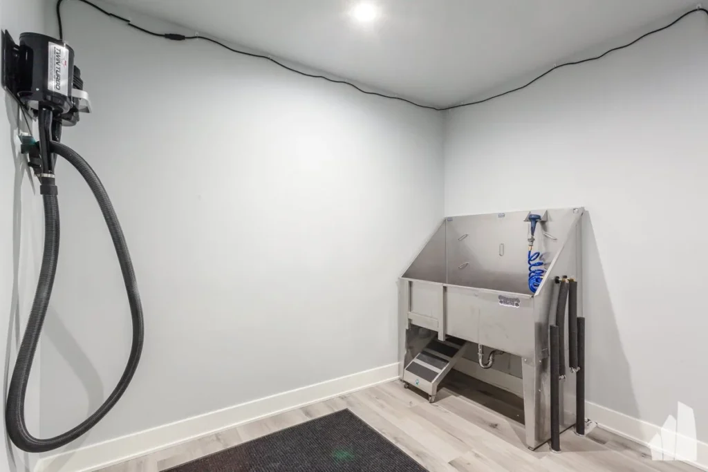 A small room with a metal pet washing station, steps, hose, and a wall-mounted grooming dryer on white walls and light wood floors.