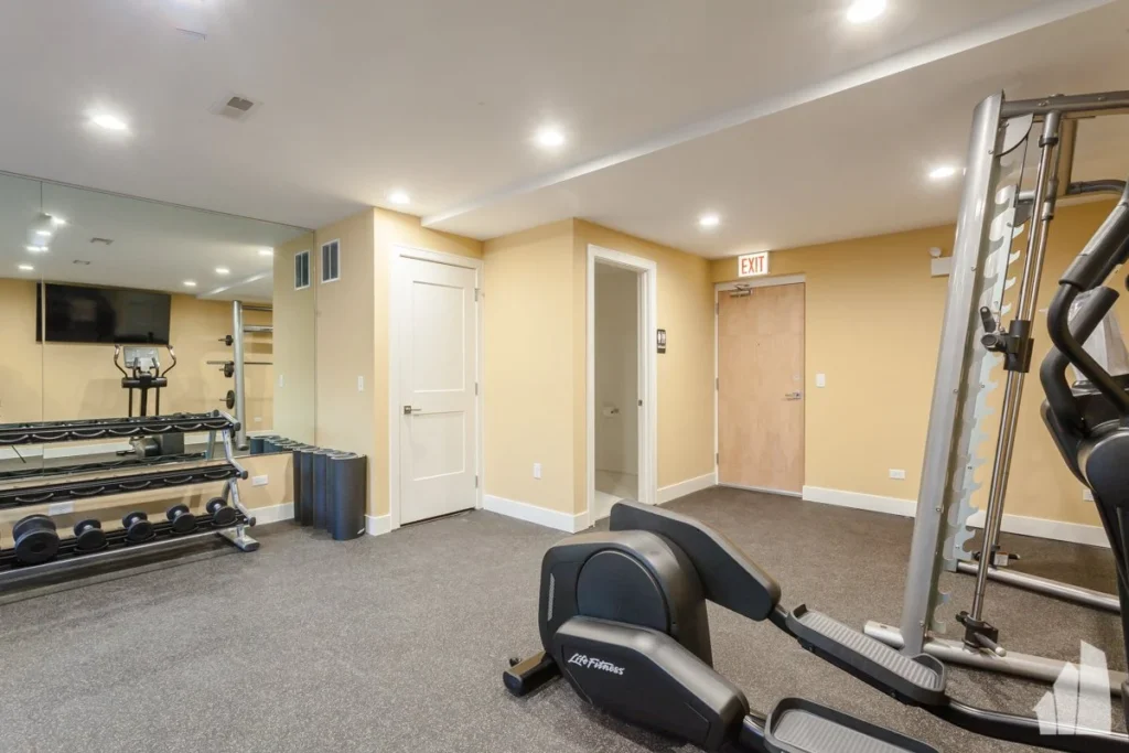 Small gym room with dumbbells, a mirror, exercise bike, strength training machine, and beige walls with overhead lighting.