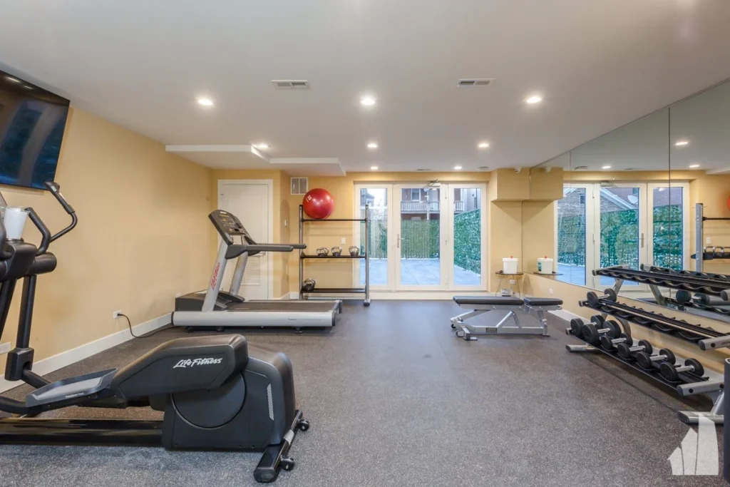 A home gym with a treadmill, weight bench, dumbbells, mirrors, a mounted TV, a red exercise ball, and large windows facing outside.