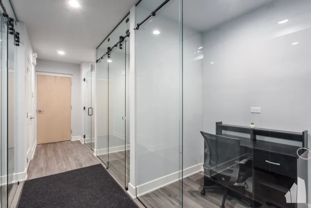 A modern hallway with glass office partitions, a closed wooden door at the end, and a small desk with a chair inside one office.