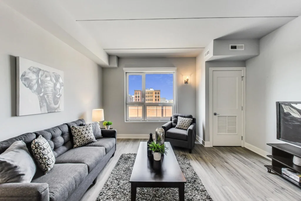 Modern living room with gray furniture, wall art of an elephant, a coffee table with plants, a window showing city buildings, and a TV on a stand.