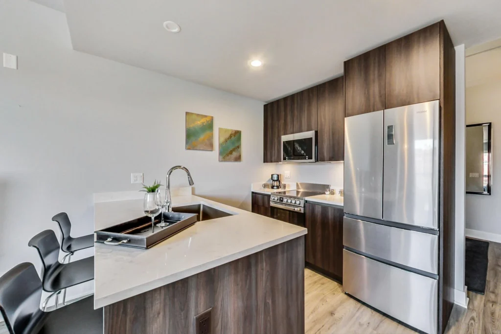 Modern kitchen with dark wood cabinets, stainless steel appliances, a white countertop, bar stools, and wall art above the sink area.