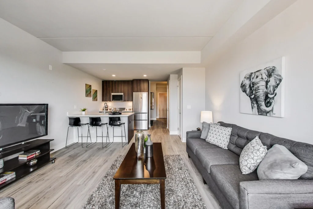 Modern open-concept living room and kitchen with gray sofa, elephant artwork, TV, breakfast bar with stools, stainless steel appliances, and wooden floors.