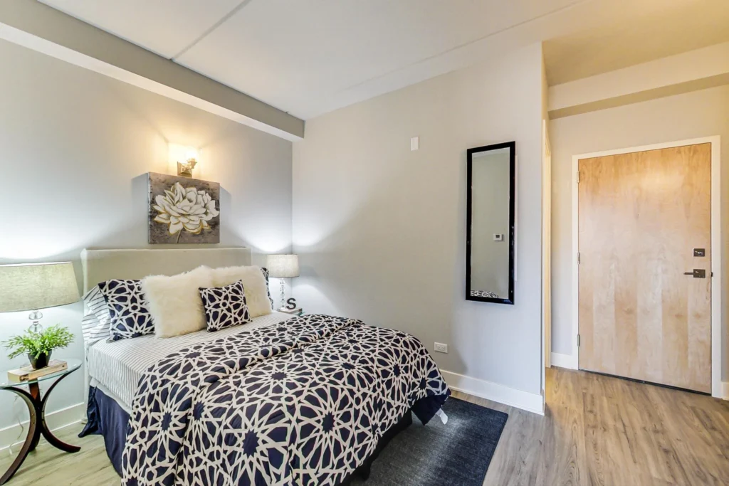 A modern bedroom with a double bed, patterned bedding, two lamps on side tables, a wall-mounted flower art piece, a full-length mirror, and a wooden door on light wood flooring.