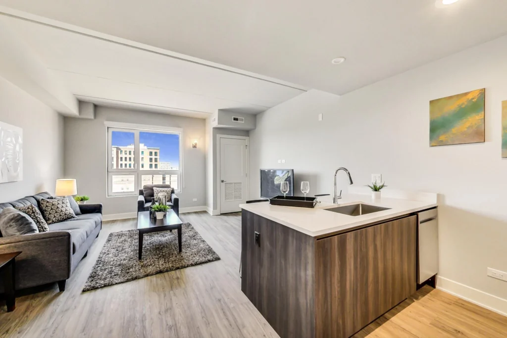 Modern open-plan apartment with a kitchen island, living area, gray sofa, coffee table, large window, and light wood floors. A painting hangs on the wall near the kitchen.