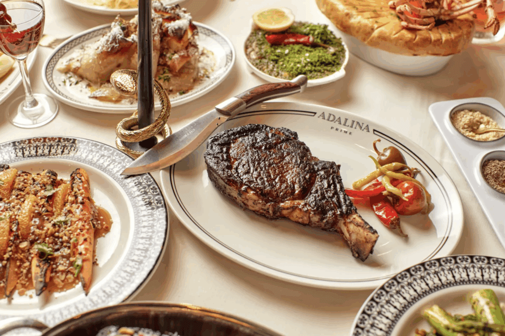 A cooked bone-in steak on a plate labeled “Adalina Prime” with peppers, surrounded by various plated dishes and a glass of red wine on a white tablecloth.