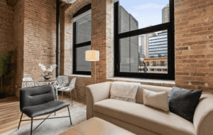 Modern loft apartment with exposed brick walls, large black-framed windows, beige sofa, black chair, and dining area with glass table and white chairs. City buildings visible outside.