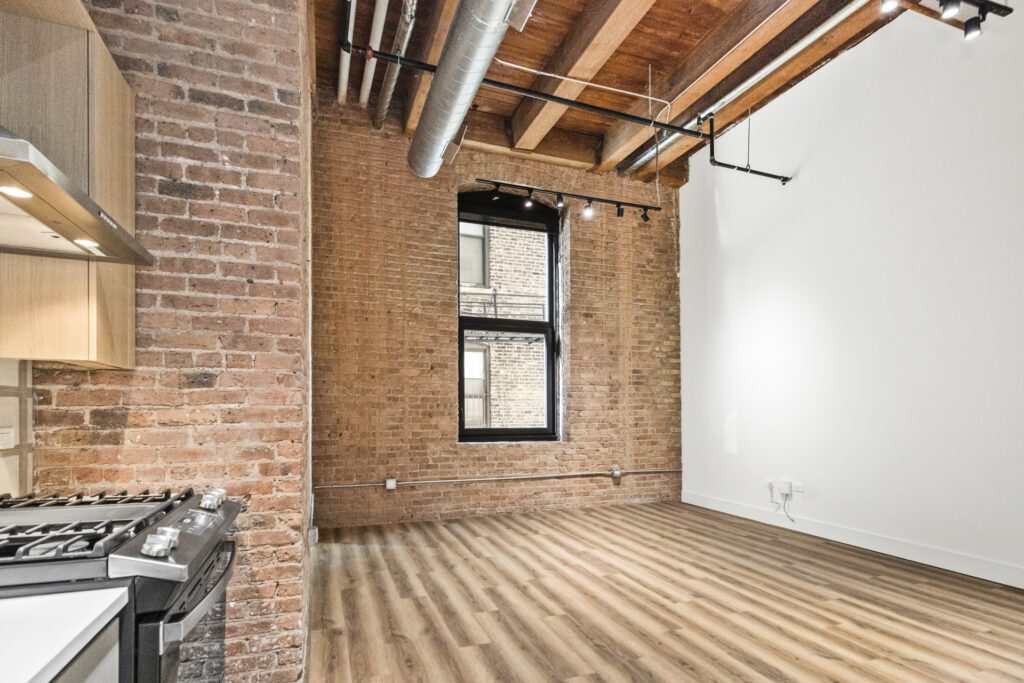 A modern loft apartment with exposed brick walls, wood flooring, a large window, and visible ceiling pipes. A kitchen stove is visible on the left.