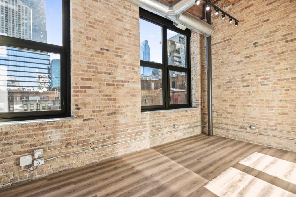 Loft-style room with exposed brick walls, large windows, hardwood floors, visible ductwork, and city buildings visible outside.