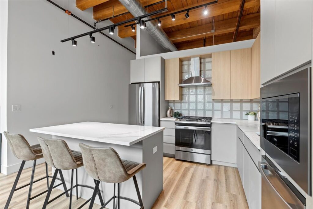 Modern kitchen with light wood floors, white cabinets, a stainless steel refrigerator, oven, and range hood, marble island with three barstools, and exposed wooden ceiling beams.