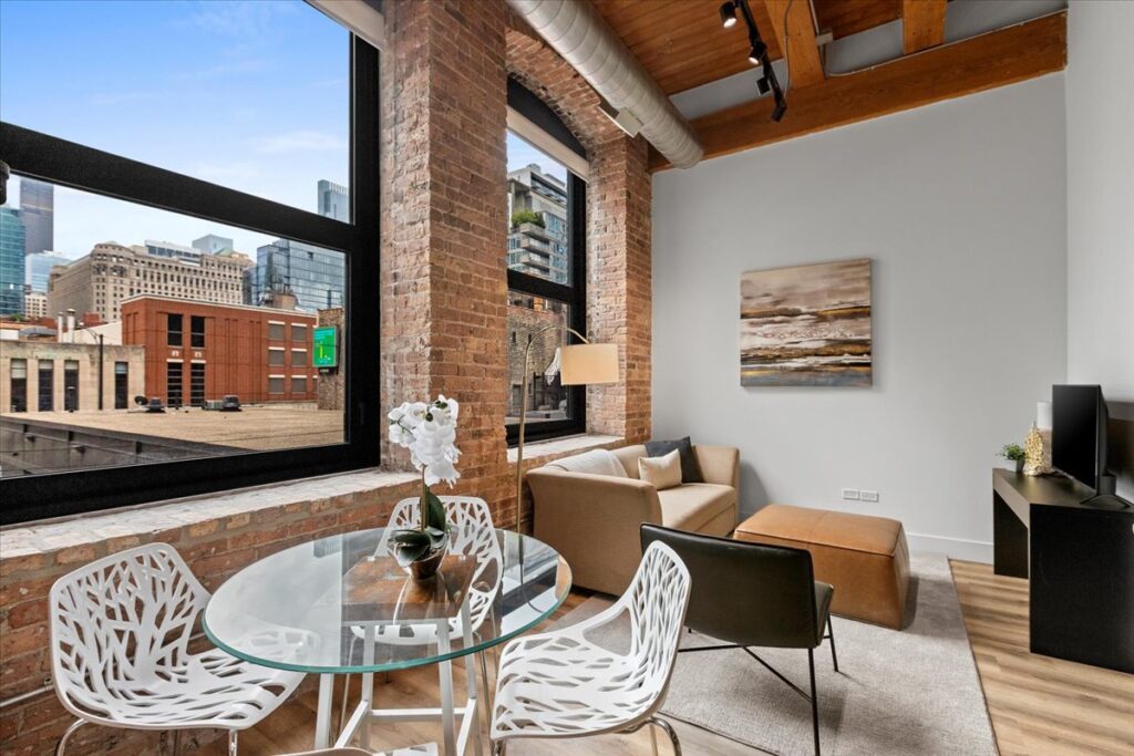 A modern loft apartment with exposed brick walls, large windows, a glass dining table with white chairs, a beige sofa, and city buildings visible outside.