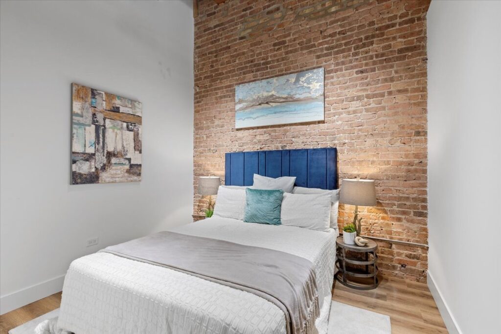 A neatly made bed with a blue headboard is centered against an exposed brick wall, flanked by two lamps and abstract artwork hanging on the walls.