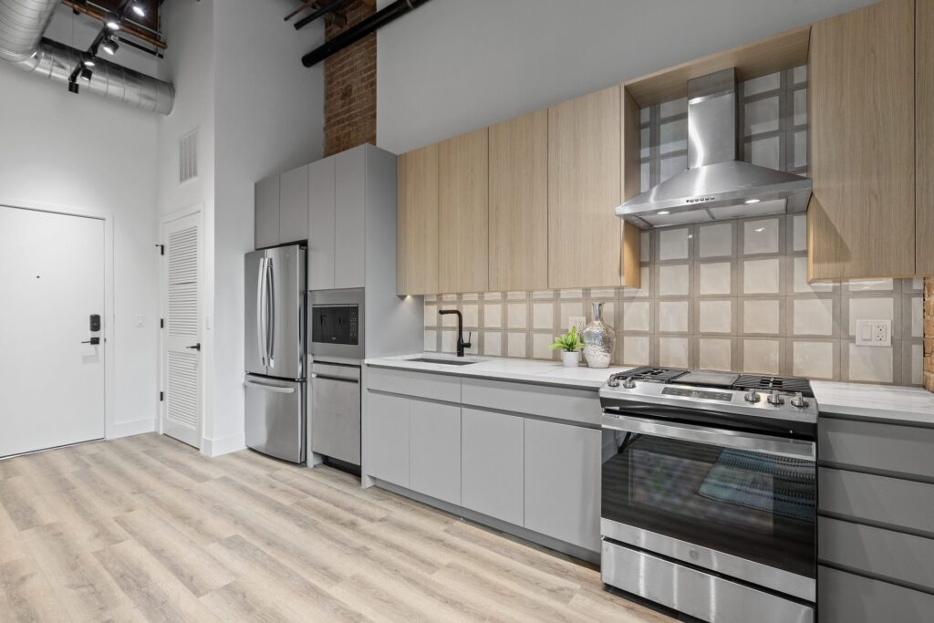 Modern kitchen with light wood and gray cabinets, stainless steel appliances, tiled backsplash, and light wood flooring.