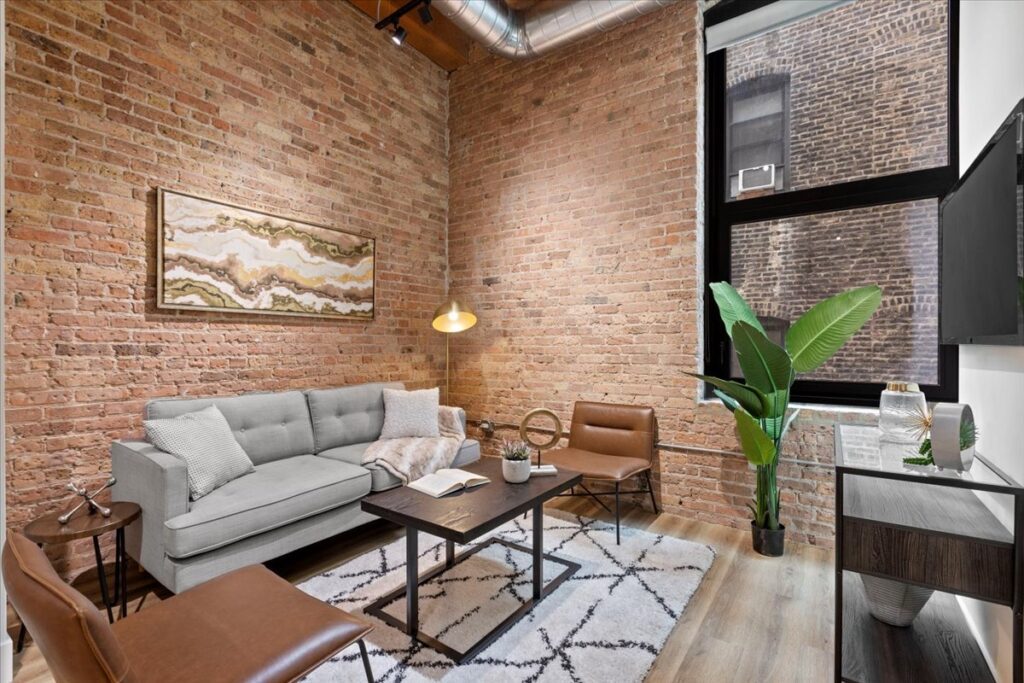 Modern living room with exposed brick walls, a gray sofa, leather chairs, a patterned rug, a wall-mounted TV, and a large window letting in natural light.