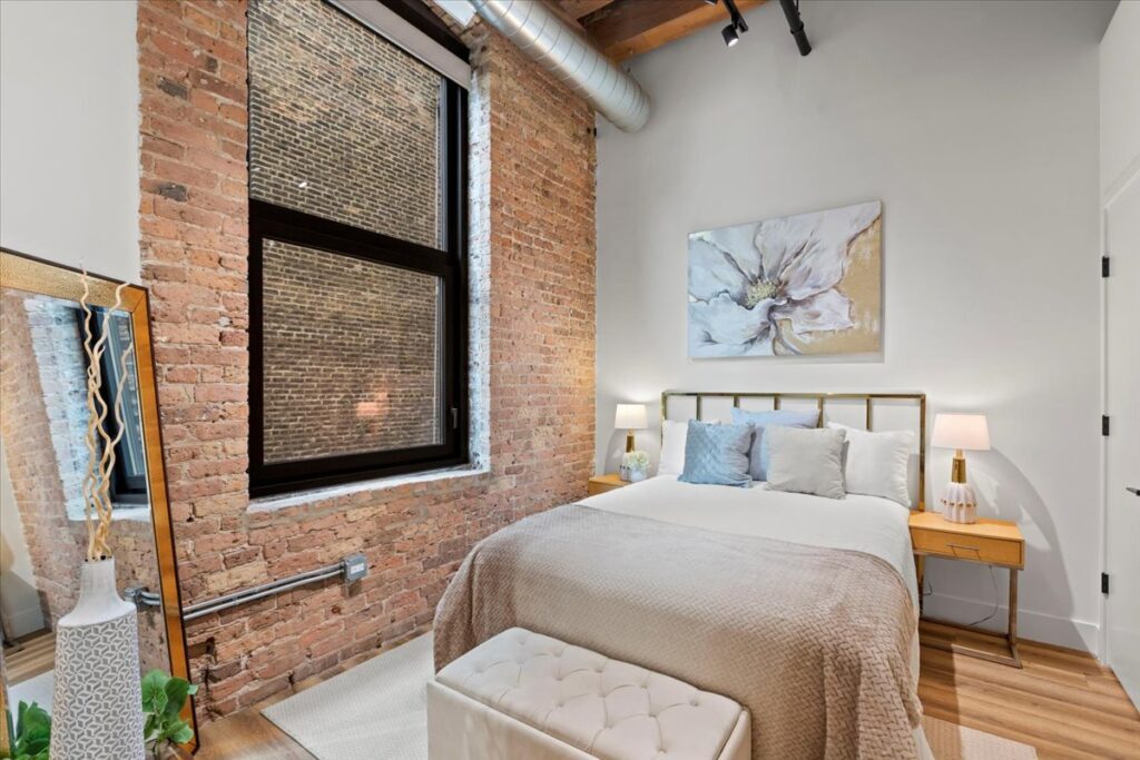 Bedroom with exposed brick walls, large window, bed with neutral bedding, two nightstands with lamps, a large flower painting, and a floor mirror.