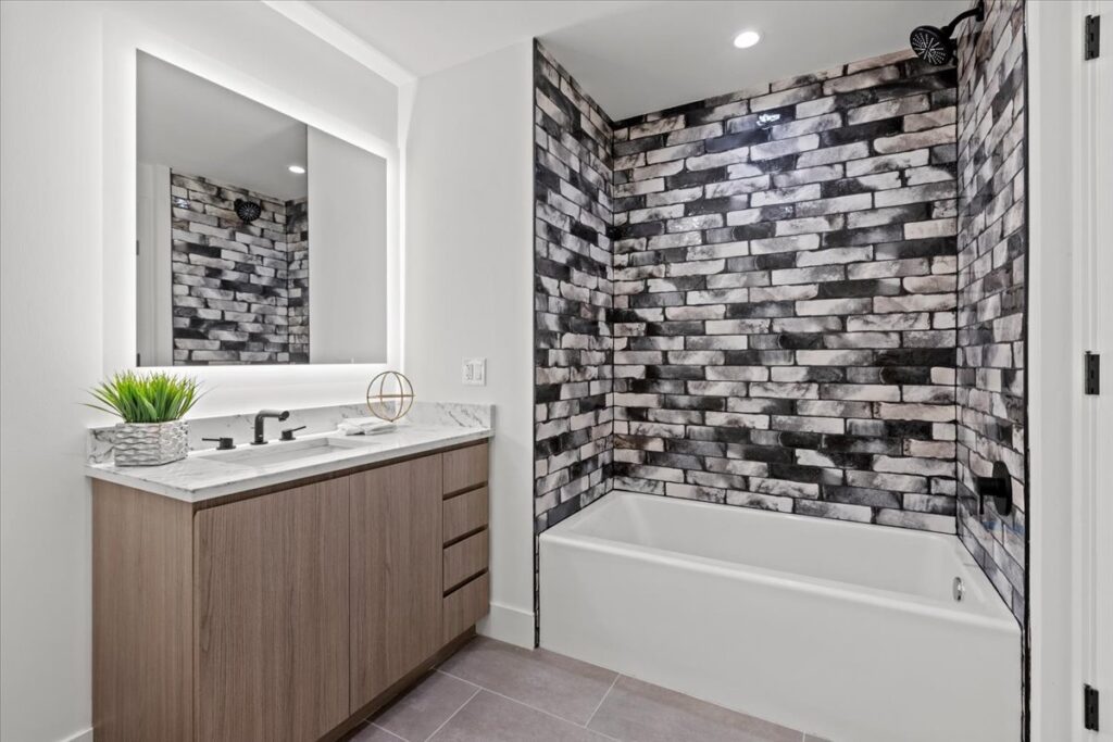Modern bathroom with a wood vanity, marble countertop, illuminated mirror, and a bathtub featuring black, white, and gray brick-patterned tiles.