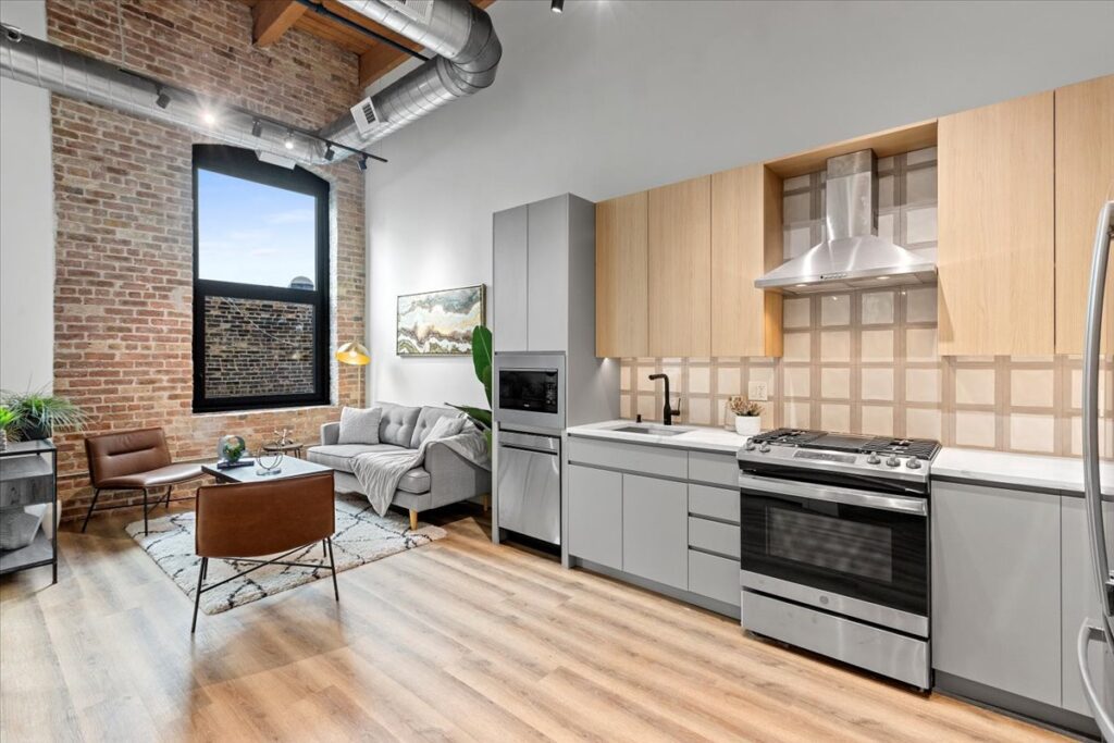 Modern loft apartment with exposed brick wall, large window, open kitchen with light wood cabinets, and a small living area with contemporary furnishings.