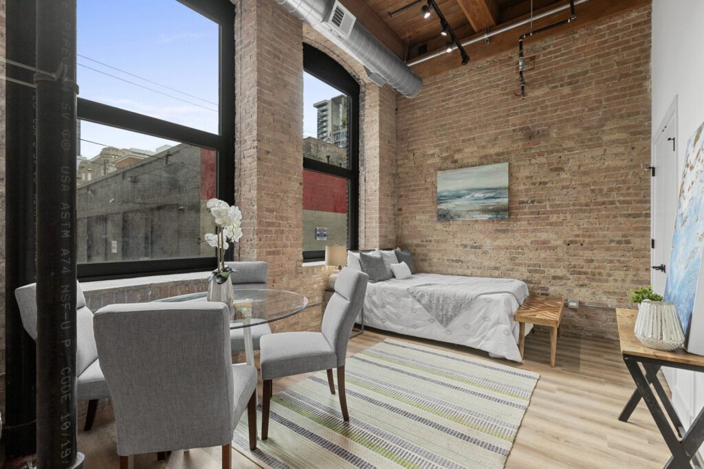 A modern loft apartment with exposed brick walls, large windows, a bed, a round glass dining table with chairs, and contemporary decor.