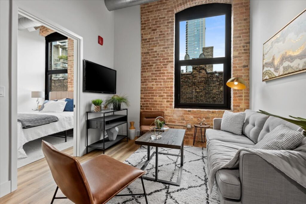 Modern living room with exposed brick walls, gray sofa, leather chair, TV, and a view into a bedroom; large window shows a cityscape outside.