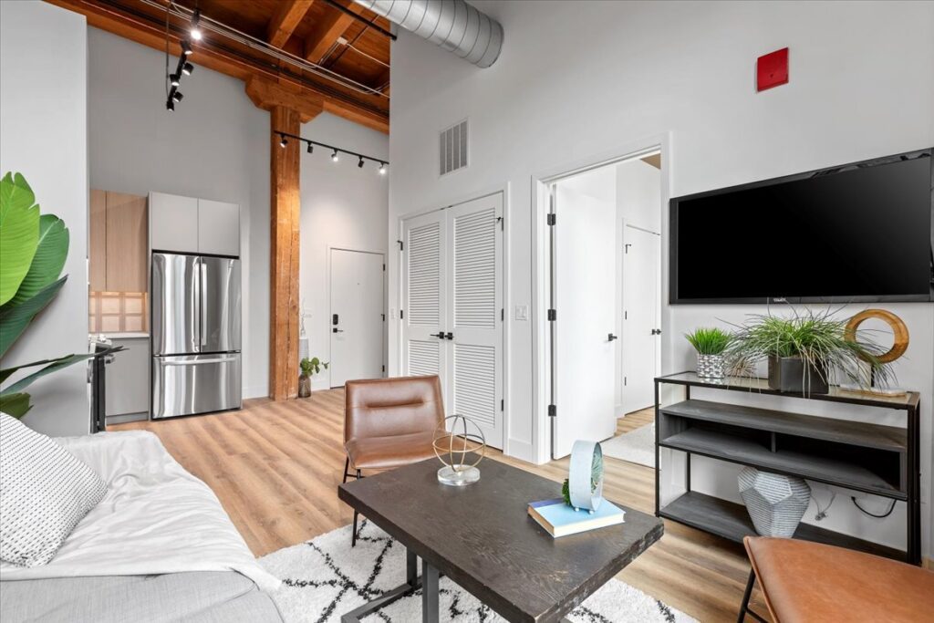 Modern apartment interior with exposed wooden beams, stainless steel kitchen appliances, a wall-mounted TV, and contemporary furniture in neutral tones.