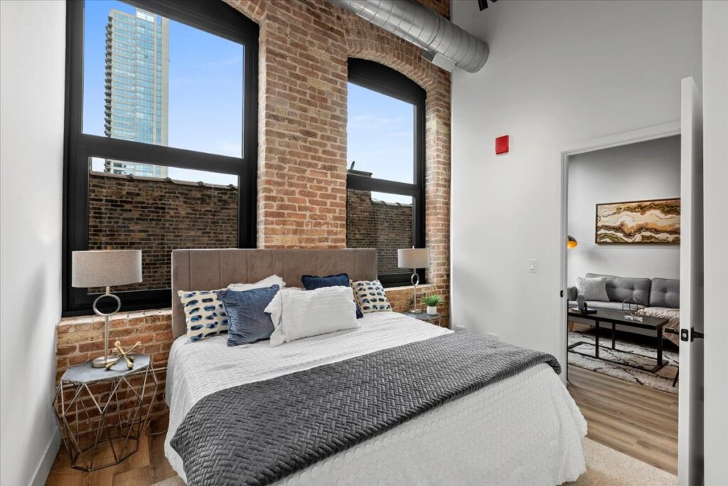 Modern bedroom with exposed brick walls, large windows, and a neatly made bed; adjoining room visible with a sofa and artwork on the wall.