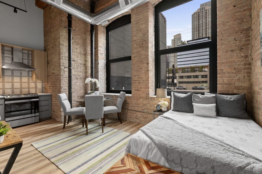 Loft apartment with exposed brick walls, large windows, a bed, a small dining table with two chairs, kitchen area, and city buildings visible outside.