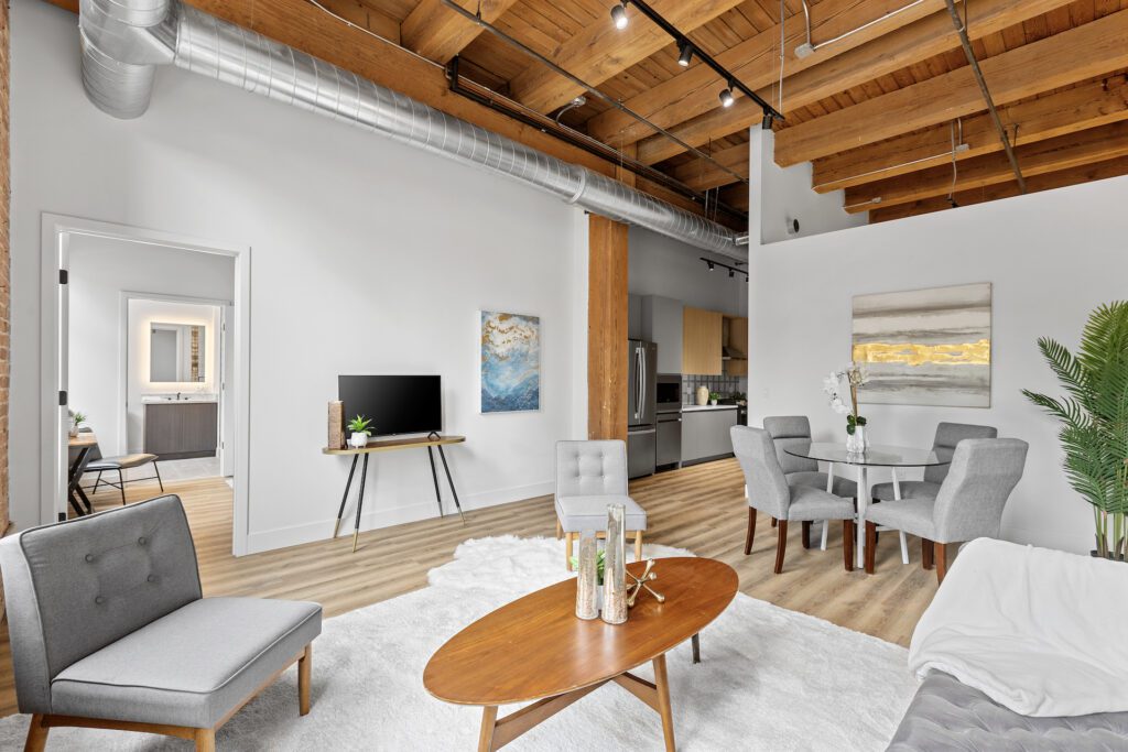 Modern loft apartment with exposed wooden beams, light wood floors, and neutral furniture. Open layout includes a living area, dining table, TV, kitchen, and visible bathroom doorway.