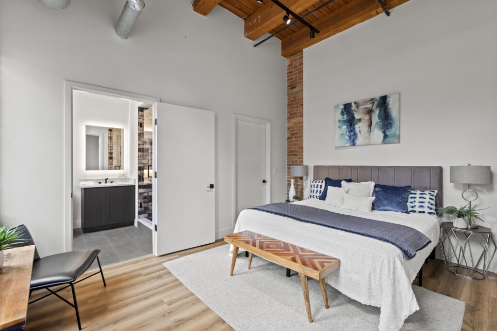 Modern bedroom with exposed brick and wood ceiling, a bed with blue and white bedding, a bench, a desk, and an open door to a bathroom.