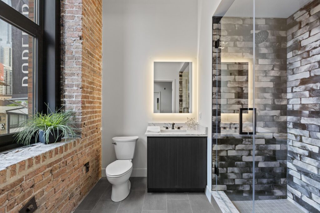 Modern bathroom with exposed brick walls, a toilet, dark vanity with illuminated mirror, glass shower, and a potted plant on the window ledge.