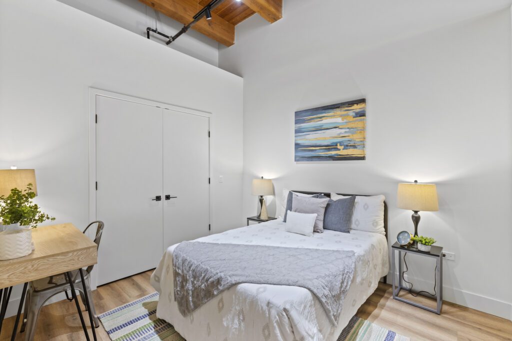 Bright bedroom with a bed, two bedside lamps, a desk with a plant, white double doors, and abstract wall art above the headboard.