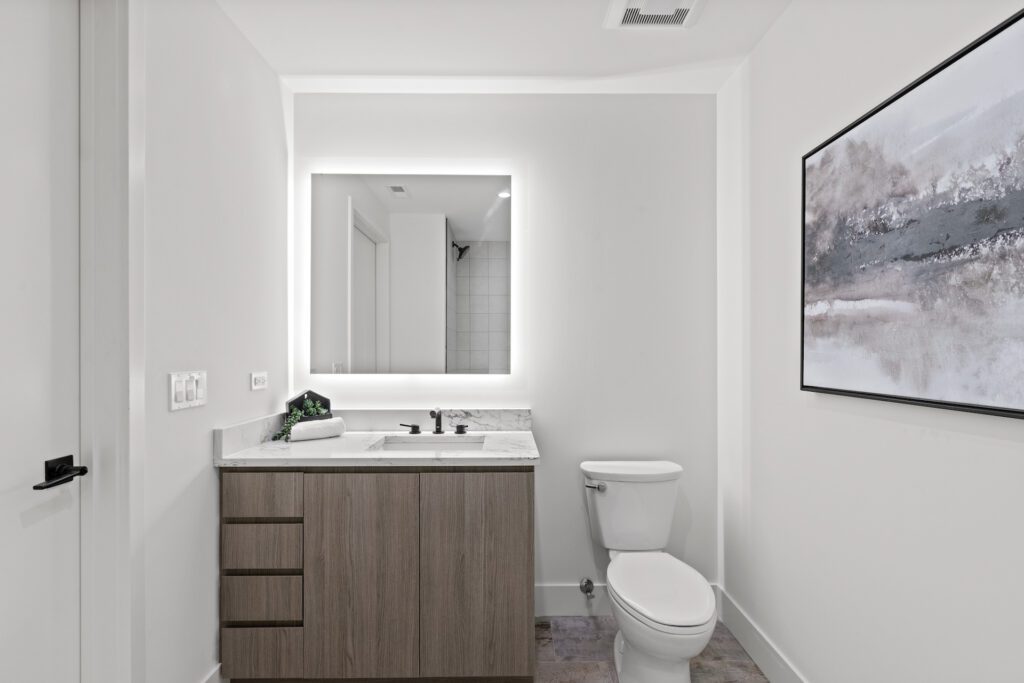 Modern bathroom with a large illuminated mirror, wooden vanity, toilet, and abstract wall art on a white wall.