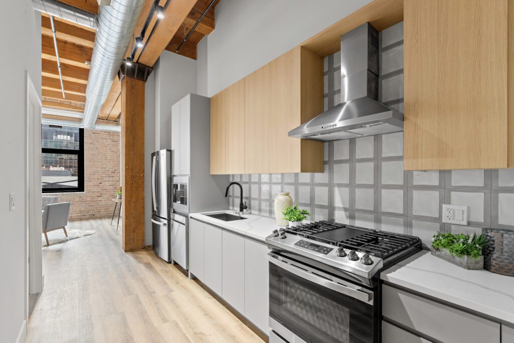 Modern kitchen with stainless steel appliances, light wood and white cabinetry, tiled backsplash, and exposed wood beams, leading to a living area with brick walls and large window.