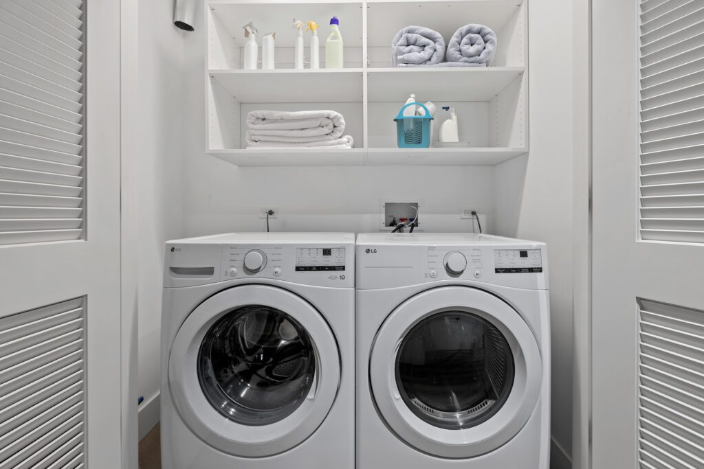 A modern laundry closet with a front-loading washer and dryer, white shelves holding towels, cleaning supplies, and detergent above the machines.