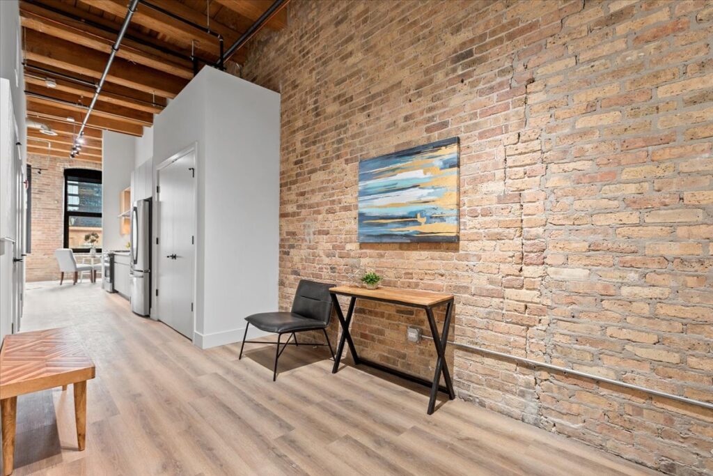 A small desk with a chair and plant sits against an exposed brick wall in a modern apartment with wooden floors and an open kitchen in the background.