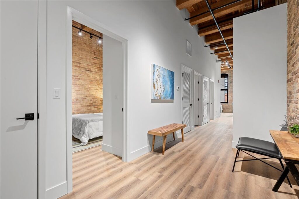 A modern hallway with light wood flooring, exposed brick and ceiling beams, a bench, wall art, and doors leading to a bedroom and other rooms.