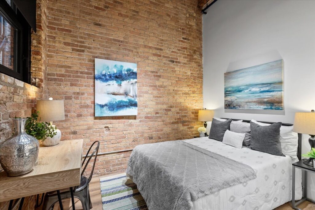 A bedroom with an exposed brick wall, a bed with gray and white bedding, two lamps, a desk with a chair, and abstract artwork on the walls.