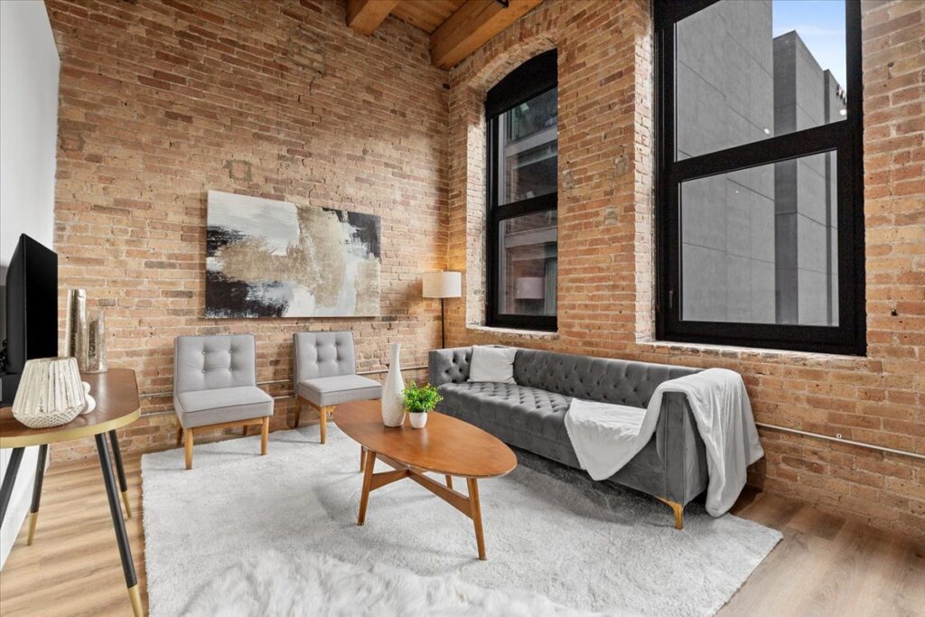 Loft-style living room with exposed brick walls, large black-framed windows, gray sofa, two gray chairs, a wooden coffee table, and neutral decor.