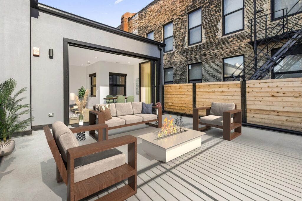 Modern outdoor patio with a fire pit, wooden lounge chairs, and a sofa, adjacent to a building with large windows and a wood fence.