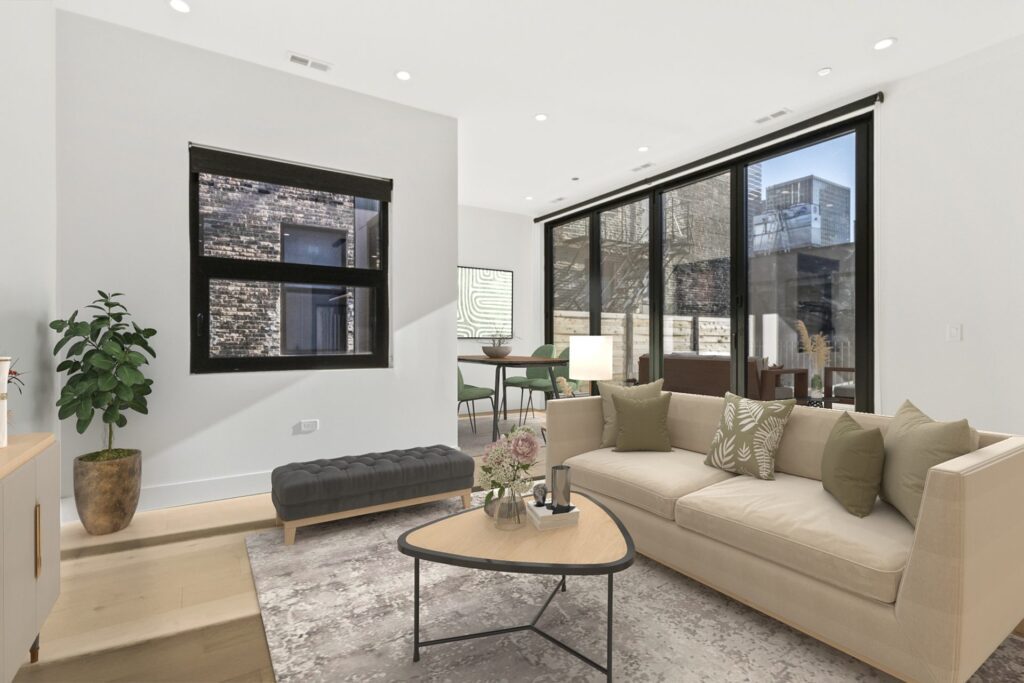 Modern living room with beige sofa, coffee table, indoor plant, and large windows showing an urban exterior; dining area visible in the background.