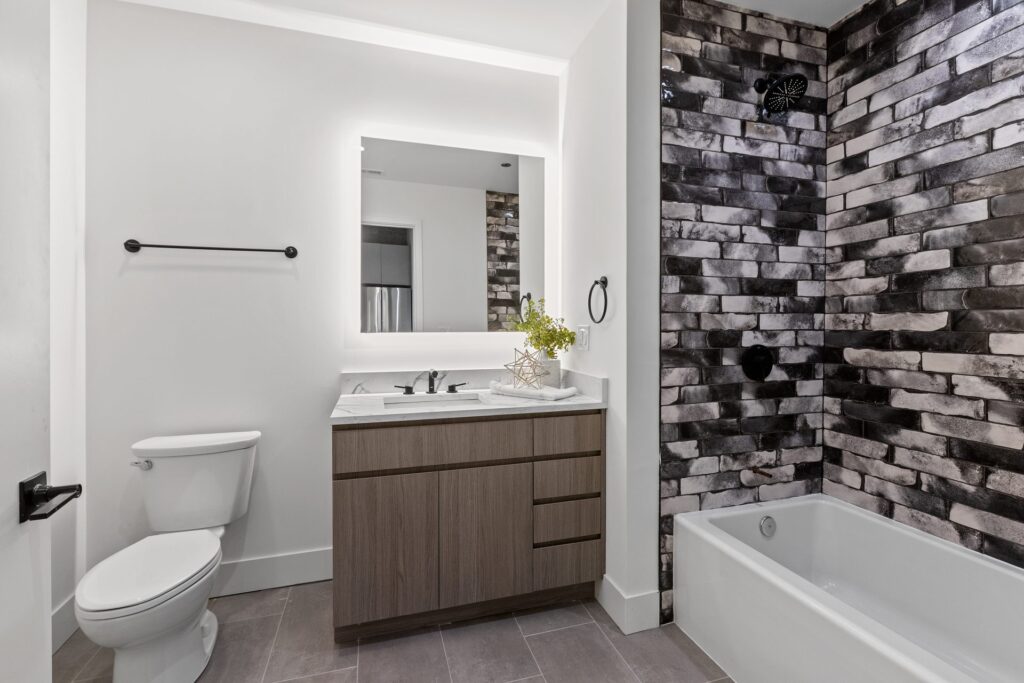 Modern bathroom with a white toilet, wood vanity, illuminated mirror, and a bathtub with black and white brick-style tile wall.