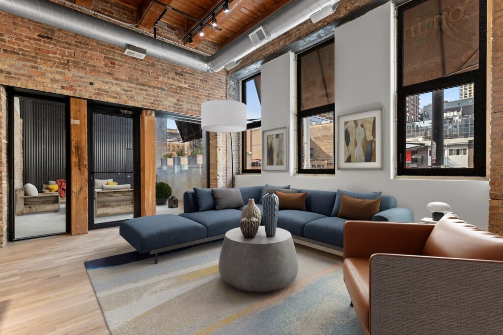 A modern living room with a blue sectional sofa, tan armchair, round coffee table, exposed brick walls, large windows, and contemporary decor.