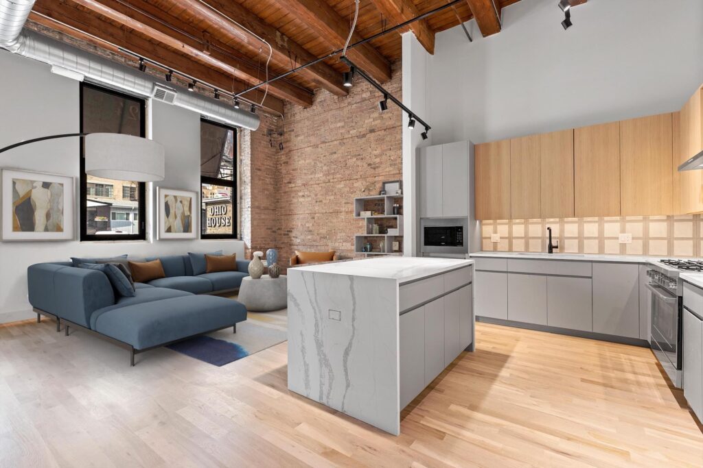 Modern open-concept kitchen and living room with exposed brick wall, wooden ceiling beams, light wood floors, and contemporary furniture.