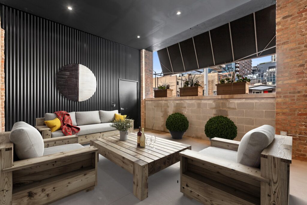 Modern outdoor patio with wooden furniture, potted plants, a decorative wall circle, and city buildings visible in the background. Bottles and glasses sit on the coffee table.